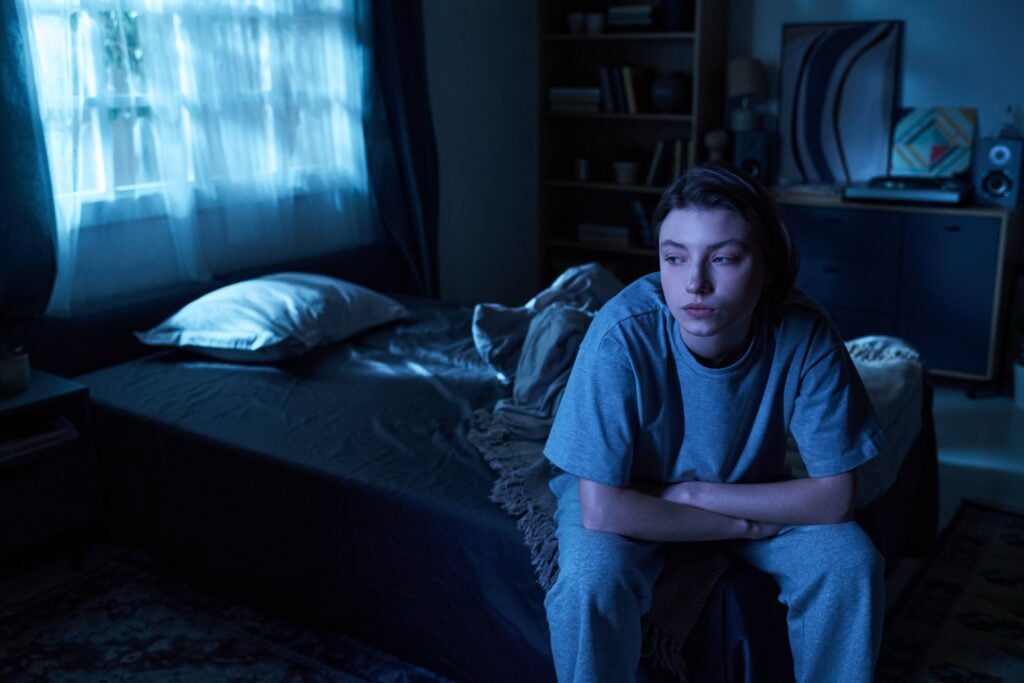 Young woman sitting on bed in dimly lit bedroom resting arms on knees looking away from camera appearing thoughtful and depressed